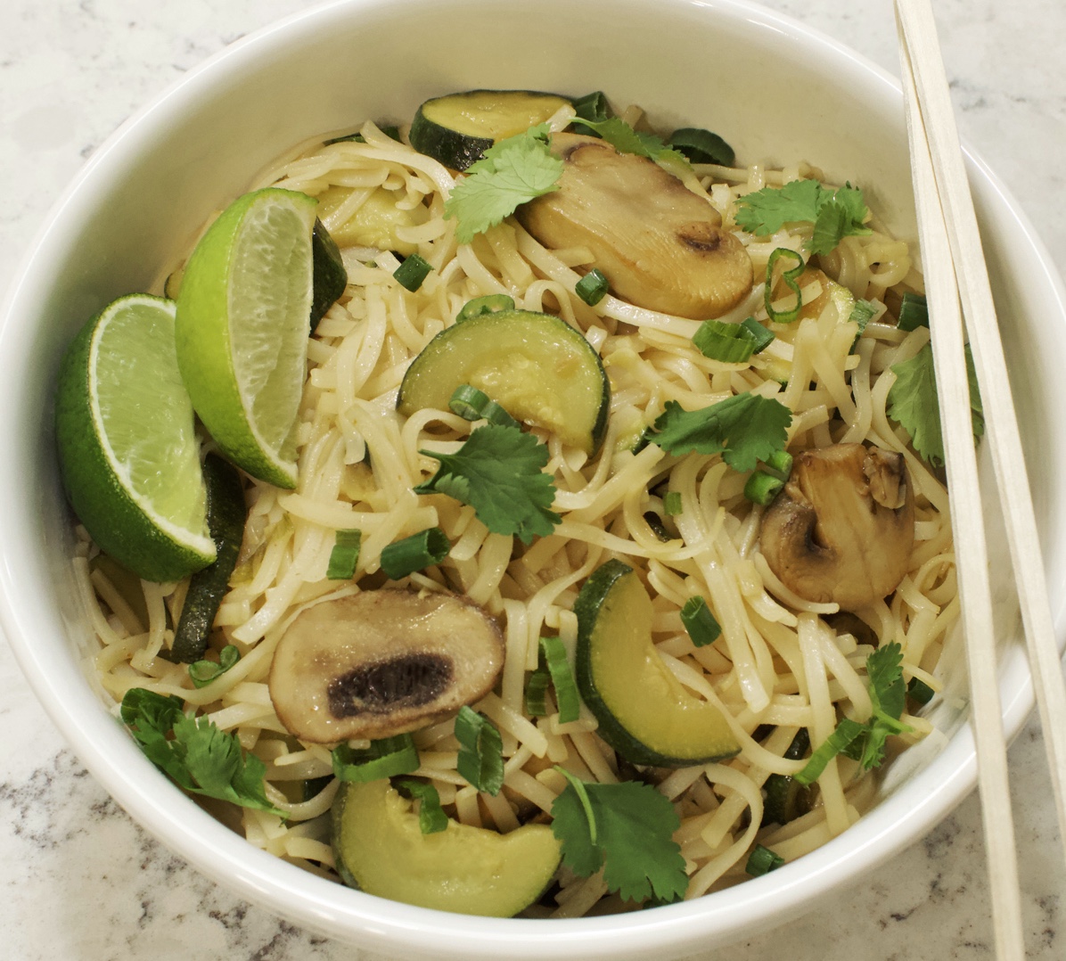 Asian style noodles in white bowl sautéed with mushrooms and zucchini. Two limes on top w chopsticks