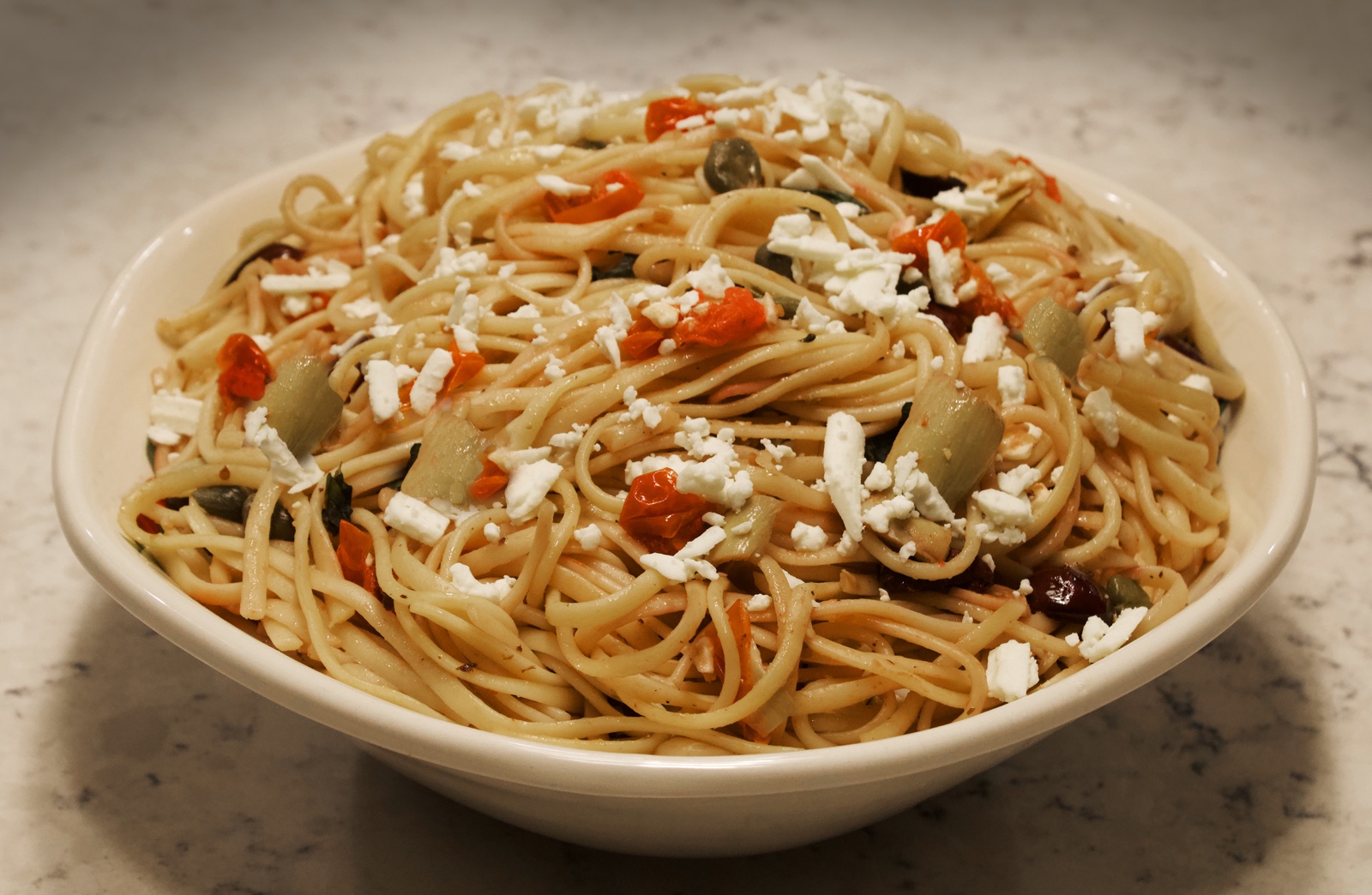 Spaghetti served in a white bowl tossed with capers, olives, tomatoes, feta cheese and artichokes
