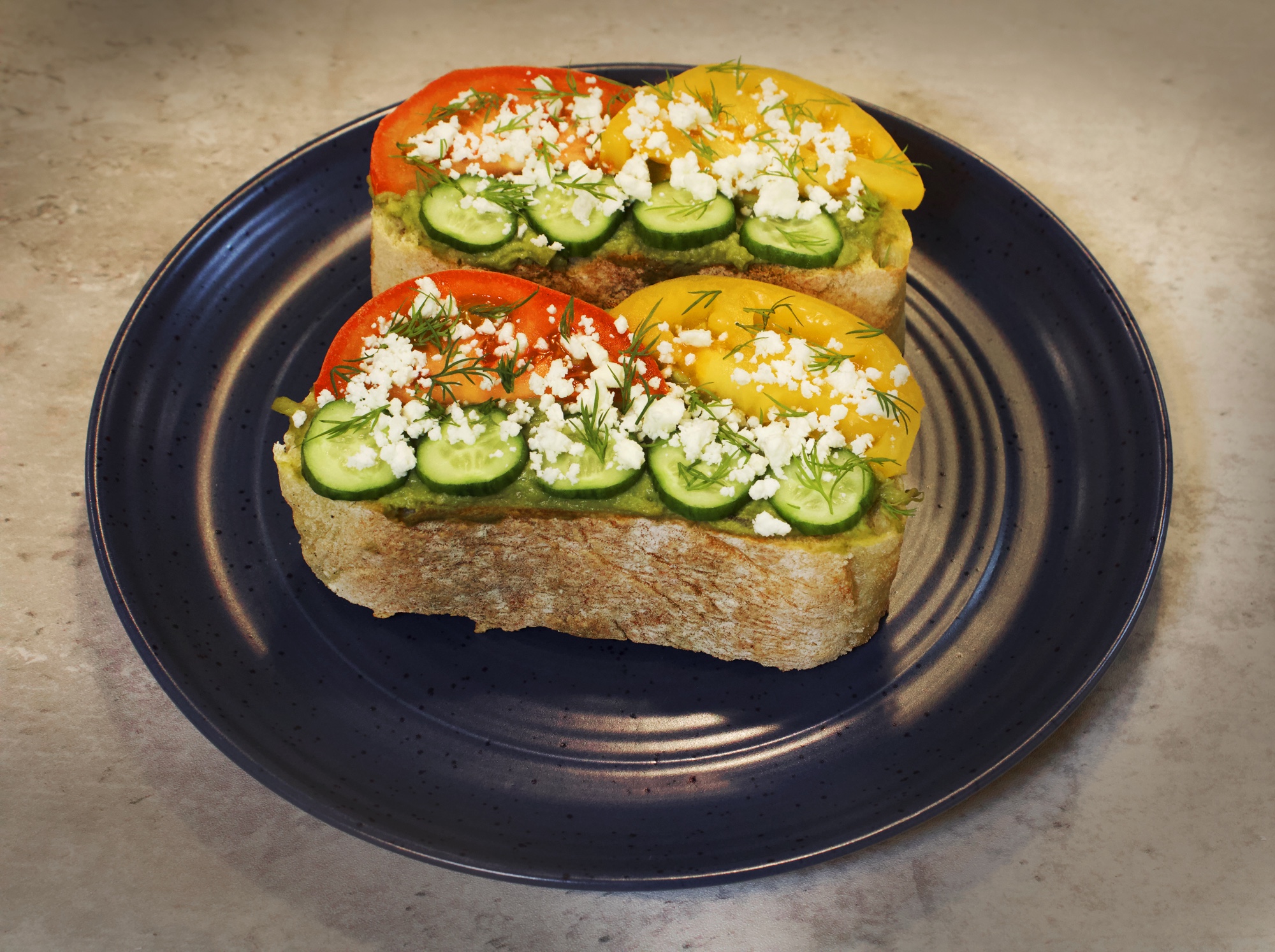 Two slices of sourdough bread with smashed avocado. Sliced tomatoes, cucumbers, feta cheese & dill. 