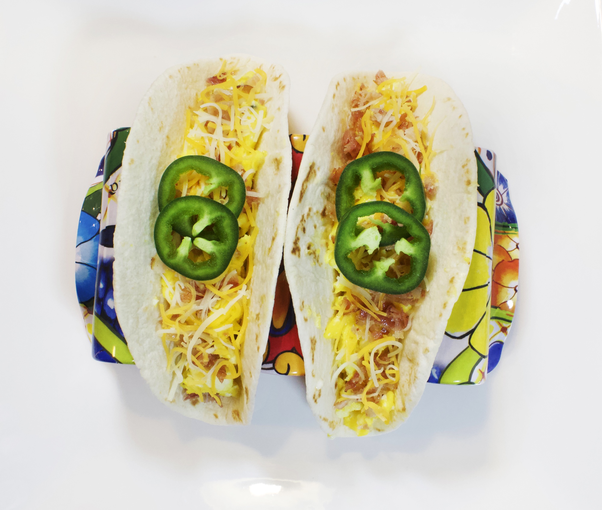 Two tacos served with scrambled eggs, pieces of bacon, shredded cheese and garnished with jalapeños.